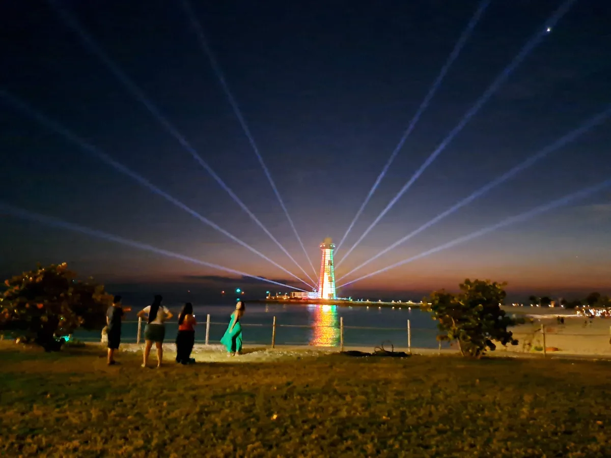 A nighttime photo shows several people on a beach looking toward a brightly lit lighthouse emitting multiple laser beams…