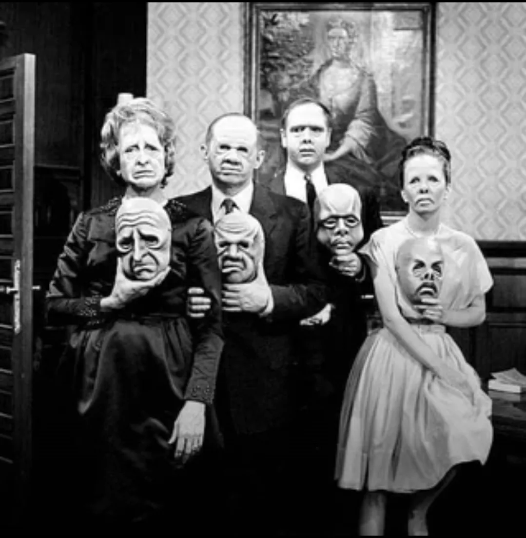 A black-and-white photo shows five people posing formally in a room, each holding a grotesque, distorted mask in front of…