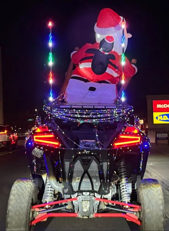 A rear view of a black ATV at night, heavily decorated with colorful string lights. On top sits a large, inflatable Santa…