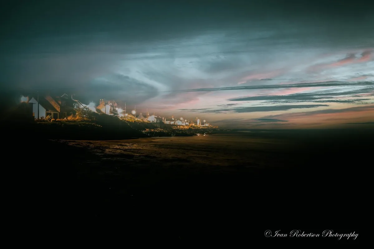A dark, atmospheric photo of a coastal village at dusk or dawn, with houses lit up on a cliff overlooking a dark beach. The…