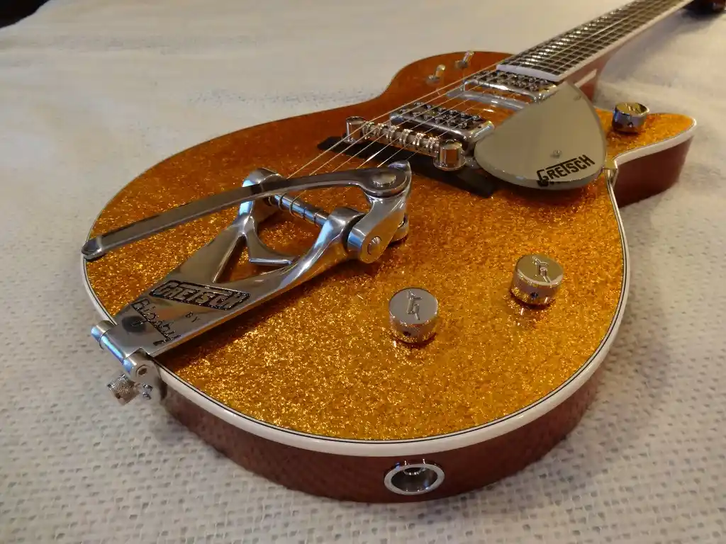 A close-up photo of a glittery gold Gretsch electric guitar with a silver tremolo arm and control knobs, resting on a white…