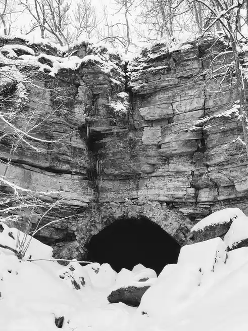 A black-and-white photo shows a snow-covered rocky cliff with a dark, arched tunnel entrance. Bare tree branches frame the…