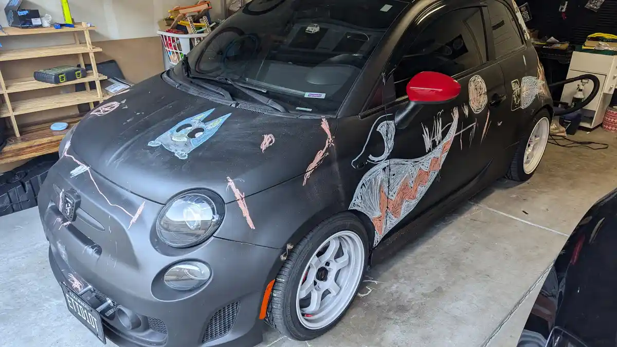 A black Fiat 500 is parked in a garage and covered in colorful chalk drawings, including a cartoon face on the hood and…