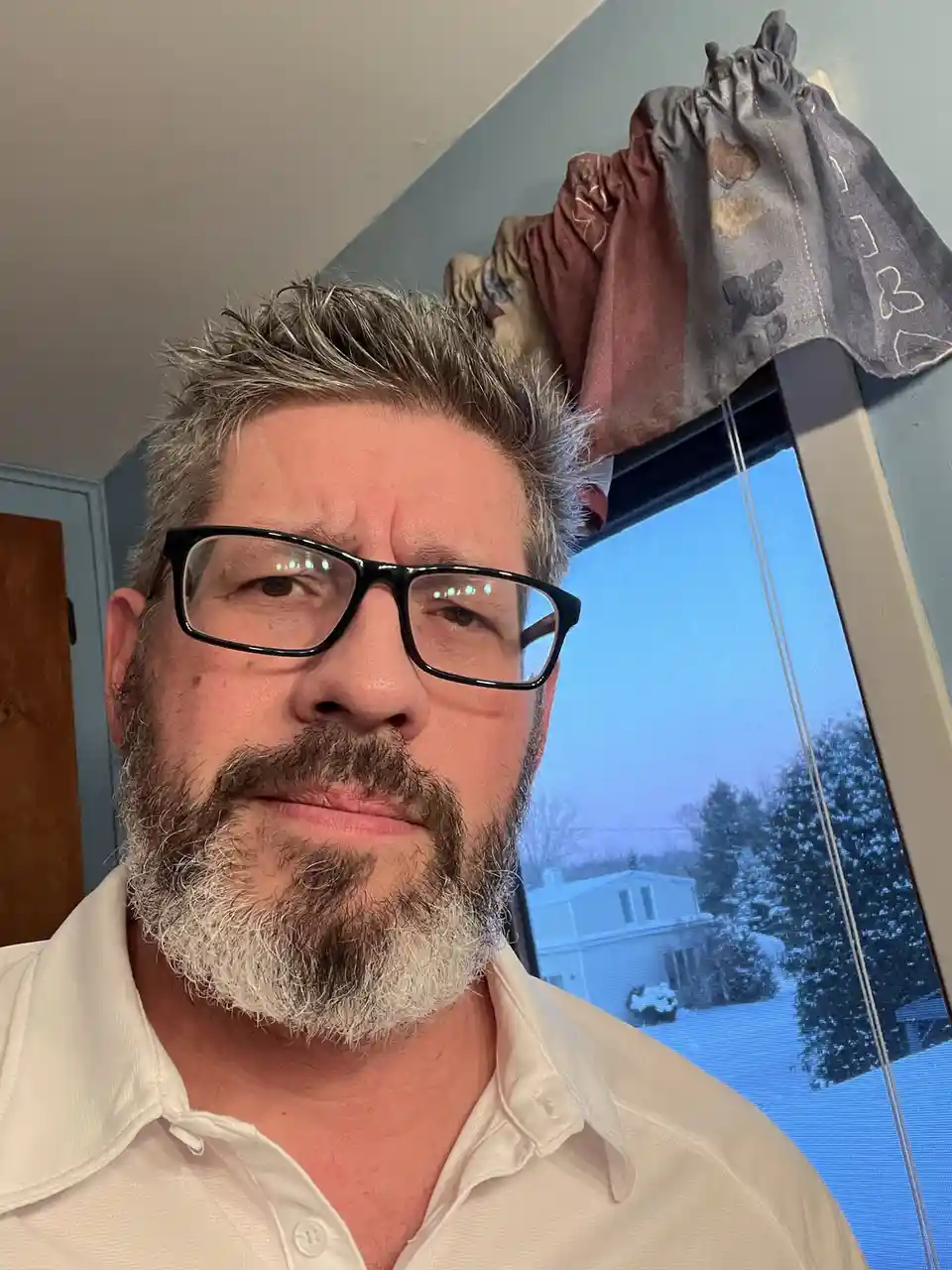 A man with glasses and a graying beard stares directly at the camera with a skeptical, unimpressed expression. Behind him, a…