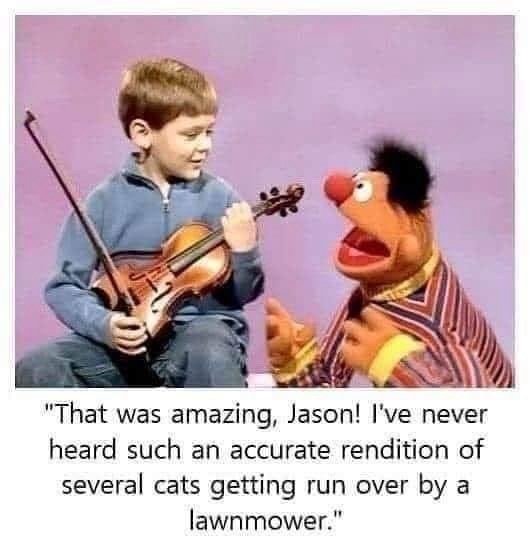 A young boy in a blue zip-up jacket holds a violin and bow while sitting next to the Muppet character Ernie, who is wearing…
