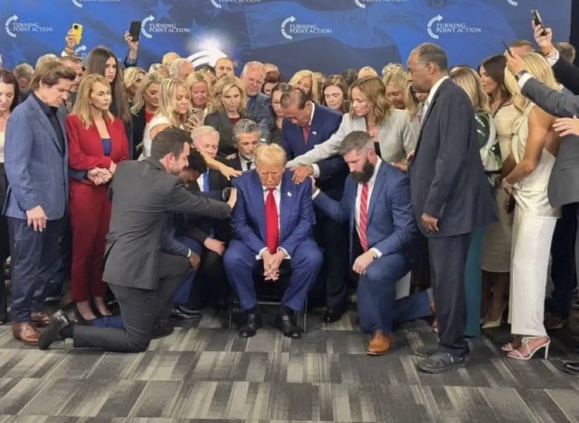 A man in a blue suit and red tie sits in a chair surrounded by a crowd of people, many of whom are kneeling or leaning in to…