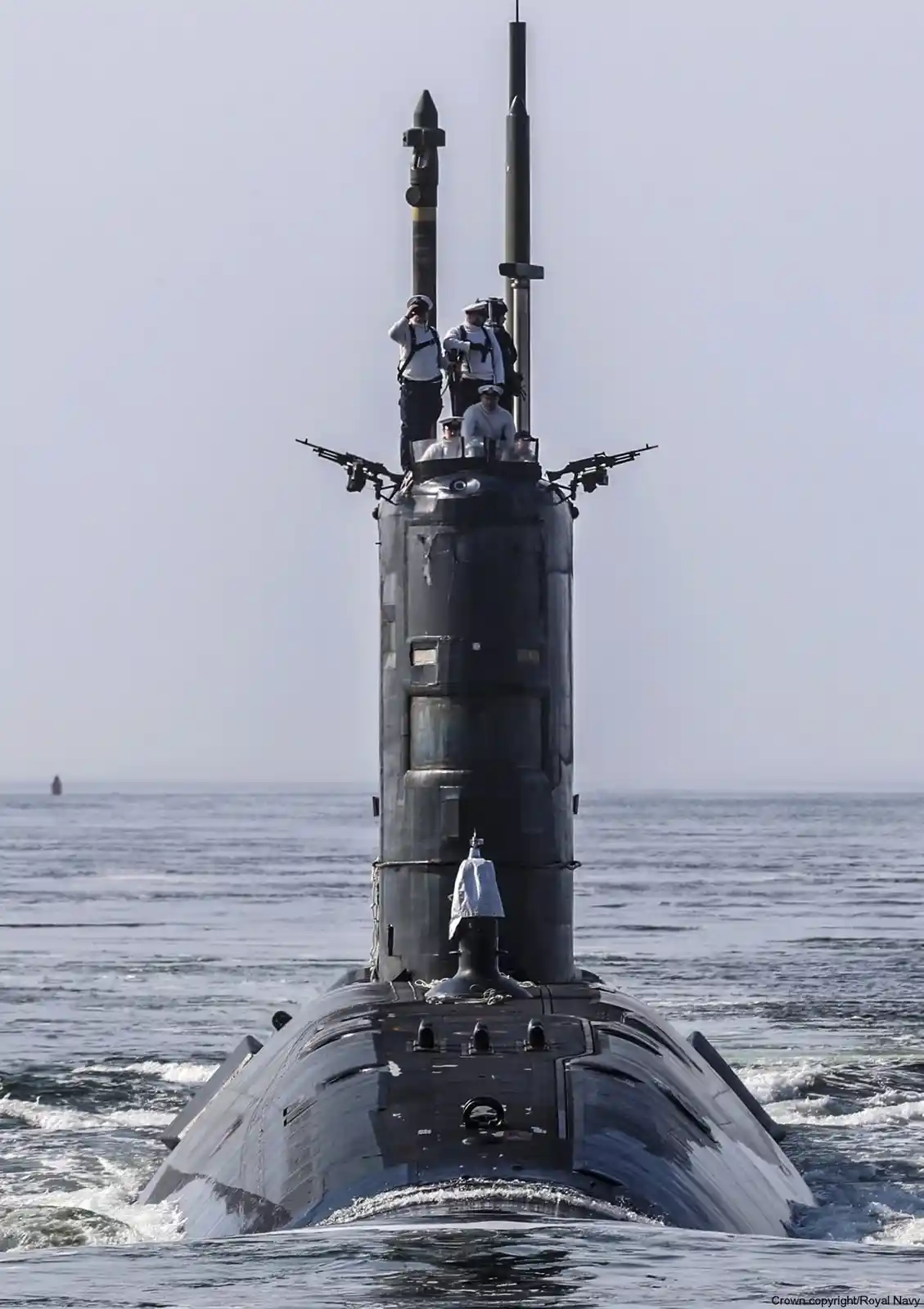 A black submarine surfaces in the ocean, its conning tower visible with several crew members in white uniforms standing atop…