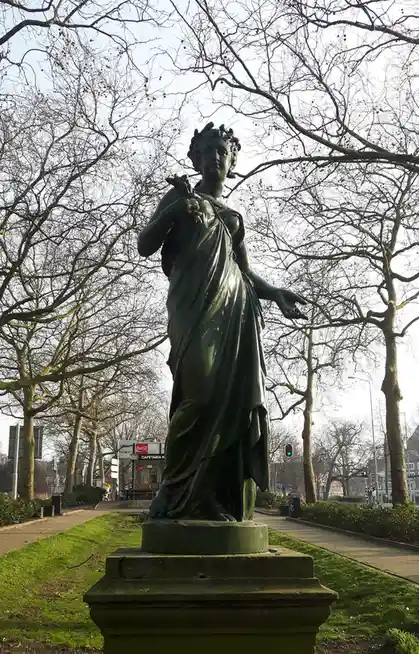 A green patina bronze statue of a classical female figure with bare breasts stands on a pedestal in a park, framed by bare…