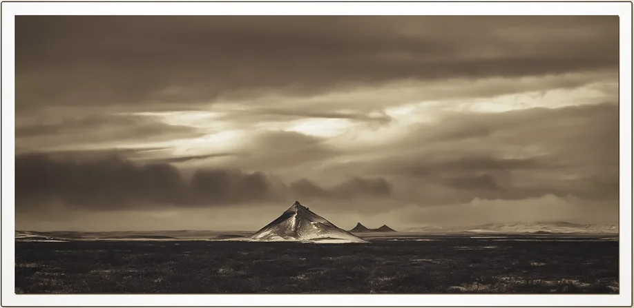 A sepia-toned photograph of a solitary, snow-capped mountain peak rising from a barren, flat landscape under a dramatic…