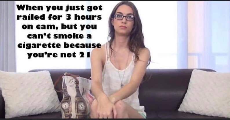 A young woman with glasses sits on a dark couch with white pillows, looking bored or annoyed. She wears a white tank top and…