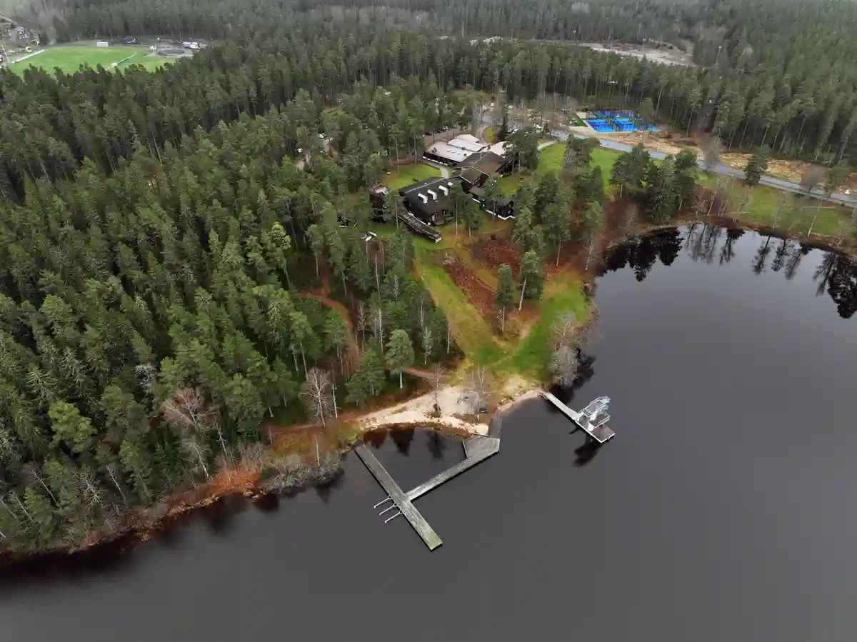 An aerial photo shows a calm lake bordered by dense green forest, with a wooden dock extending into the water and a cluster…