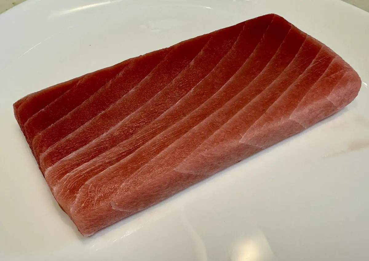 A close-up photo shows a single, thick, raw tuna steak with diagonal scoring on a plain white plate. The overlaid text reads…