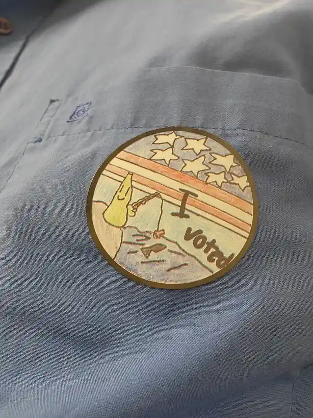 A circular 'I Voted' sticker with an American flag design is pinned to the pocket of a blue shirt. The sticker features a…