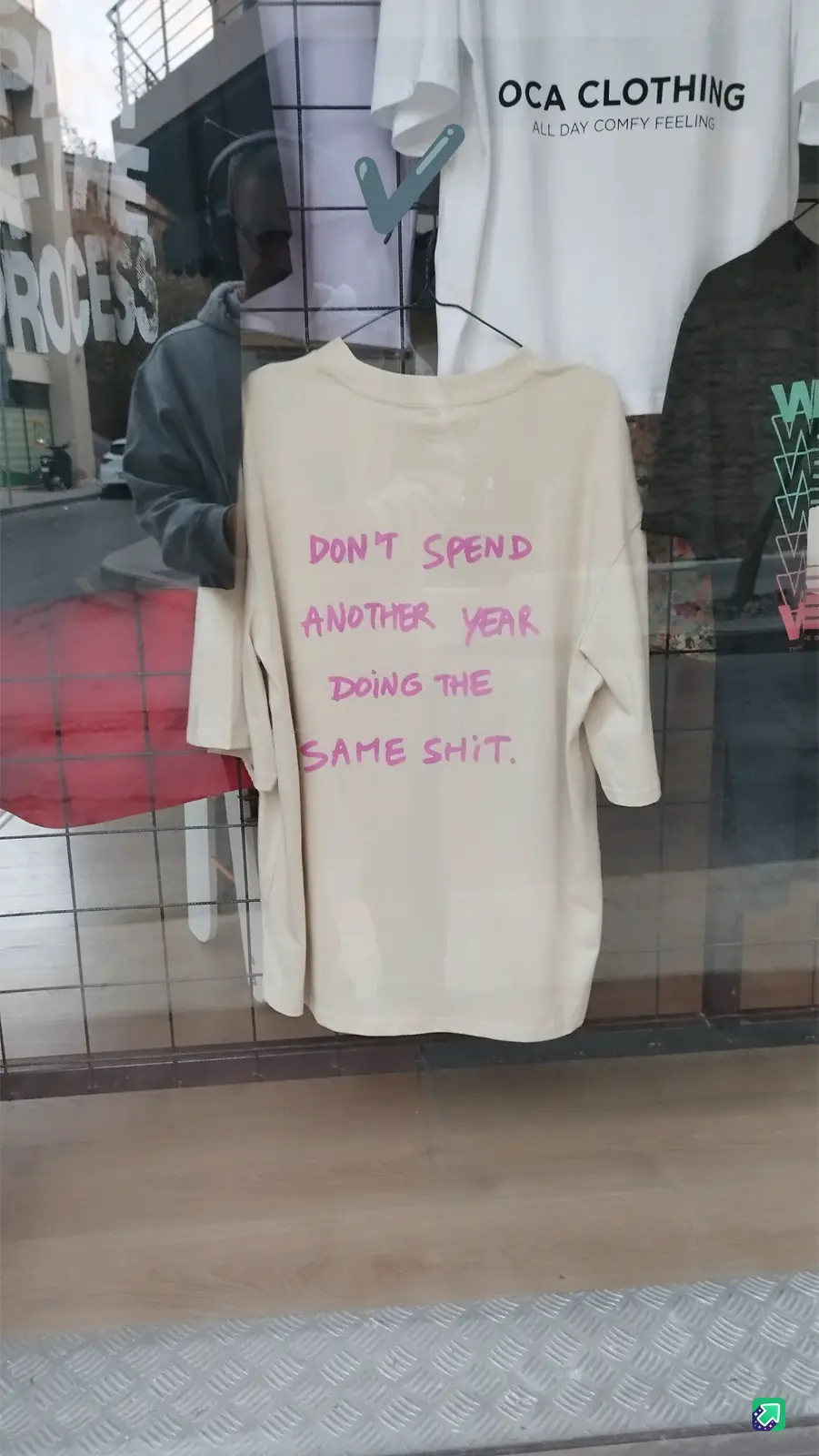 A beige t-shirt hangs in a clothing store window, displaying the handwritten pink text 'DON'T SPEND ANOTHER YEAR DOING THE…