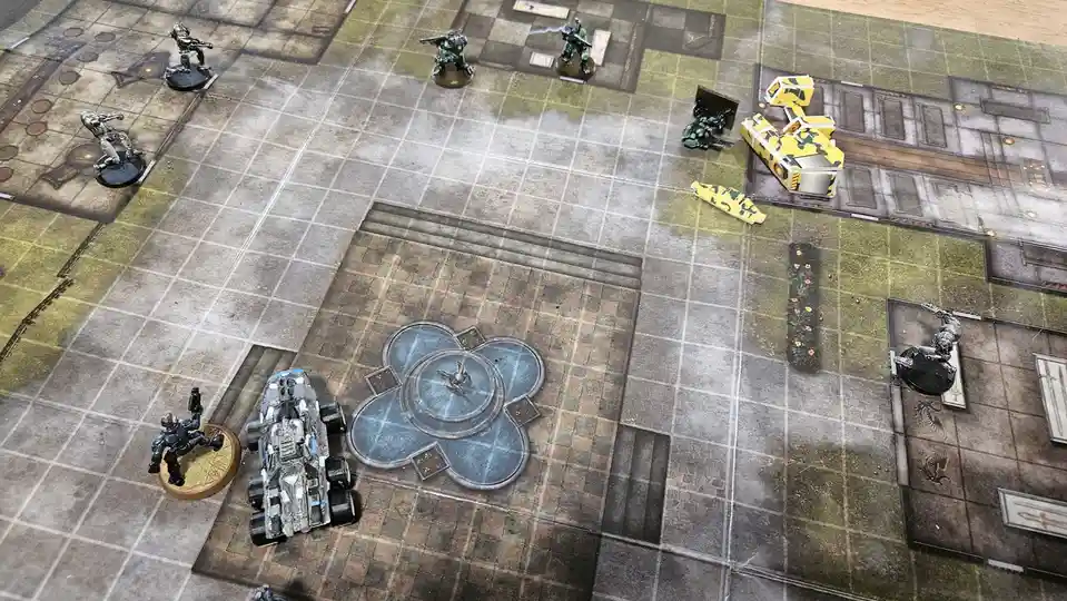An overhead shot of a tabletop wargame battlefield with miniature figures and vehicles on a printed grid map. Several…