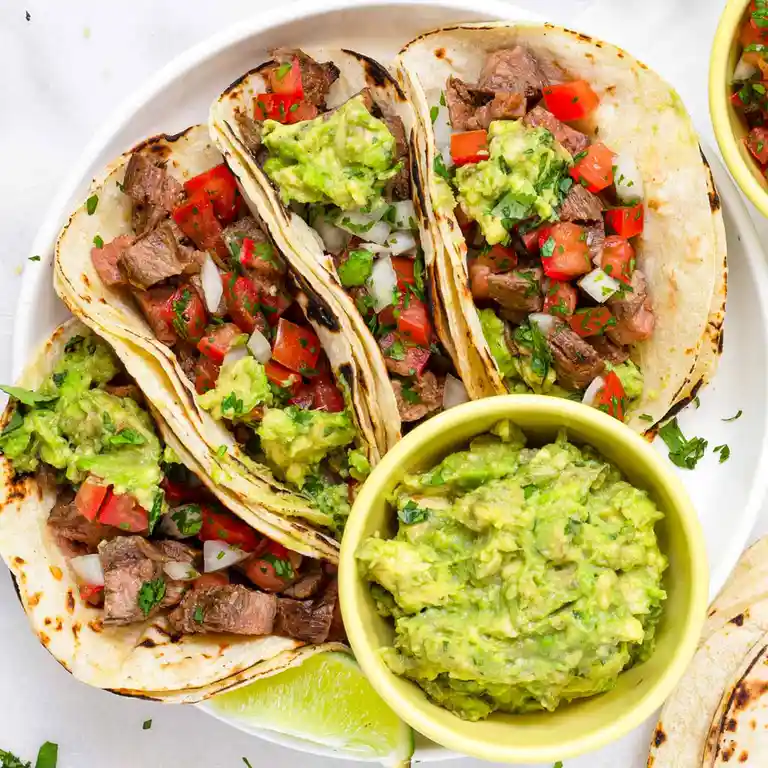 A top-down photo of four soft tacos filled with meat, diced tomatoes, onions, and guacamole, arranged on a white plate with…