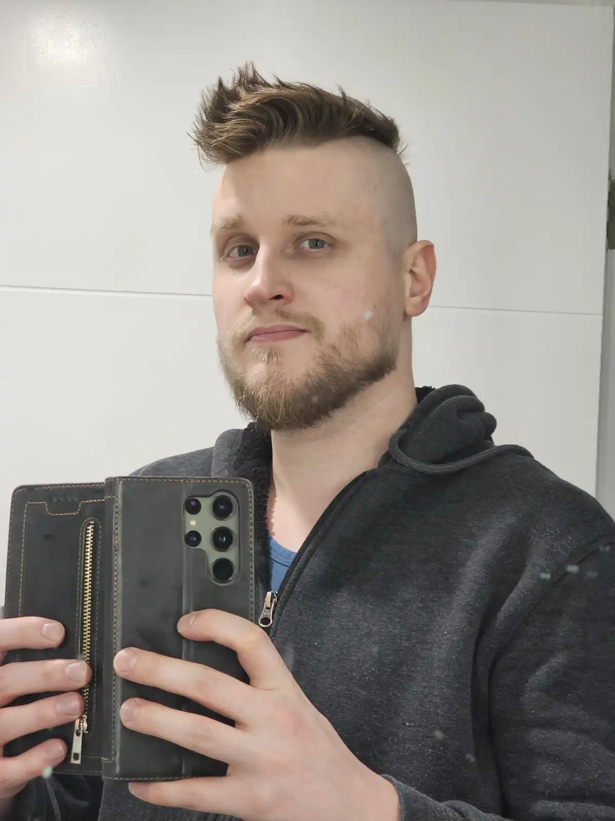 A man with a faded undercut hairstyle and a goatee takes a mirror selfie holding a smartphone in a black leather case. He…