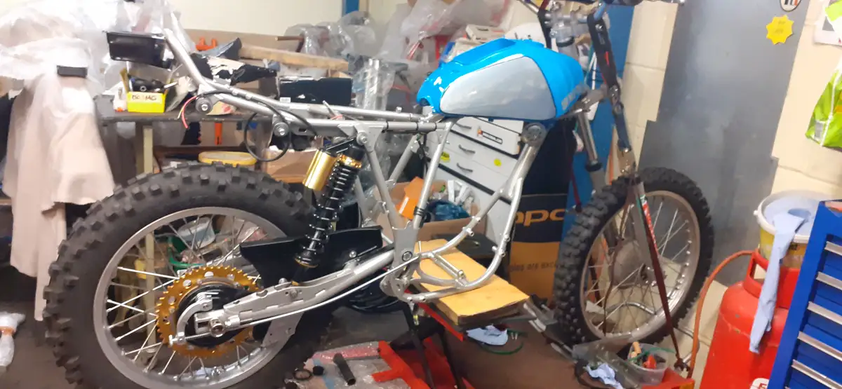 A partially assembled motorcycle with a blue fuel tank and silver frame sits in a cluttered garage, surrounded by tools and…
