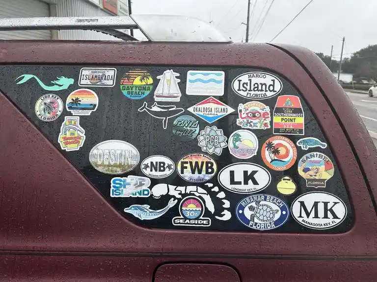 A close-up of a maroon car's rear window covered in dozens of colorful Florida-themed stickers, including 'Destin', 'Marco…