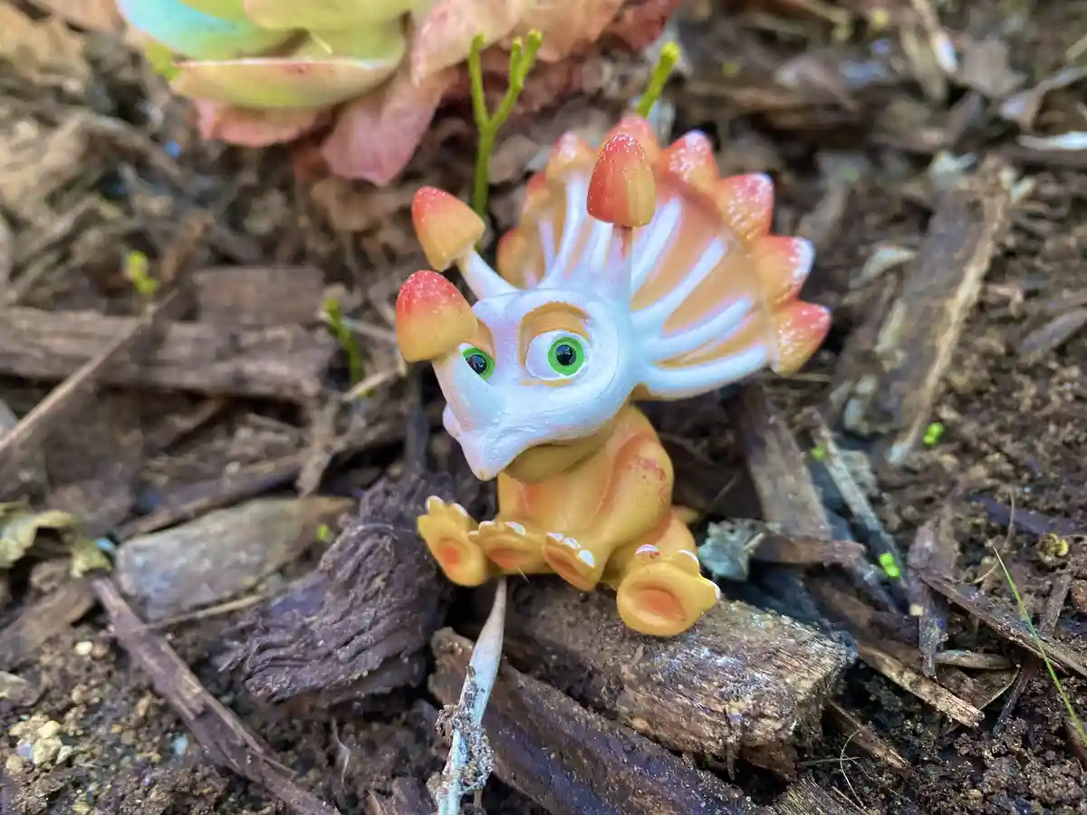A small, colorful plastic triceratops toy with large green eyes sits on a piece of wood in dirt and mulch, surrounded by…