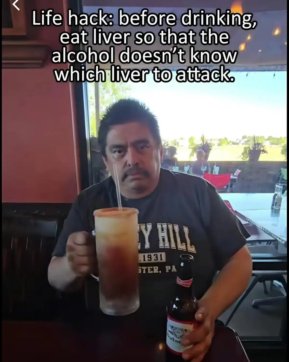 A man sits at a table in a restaurant, holding a large drink with a straw and a beer bottle, looking directly at the camera…