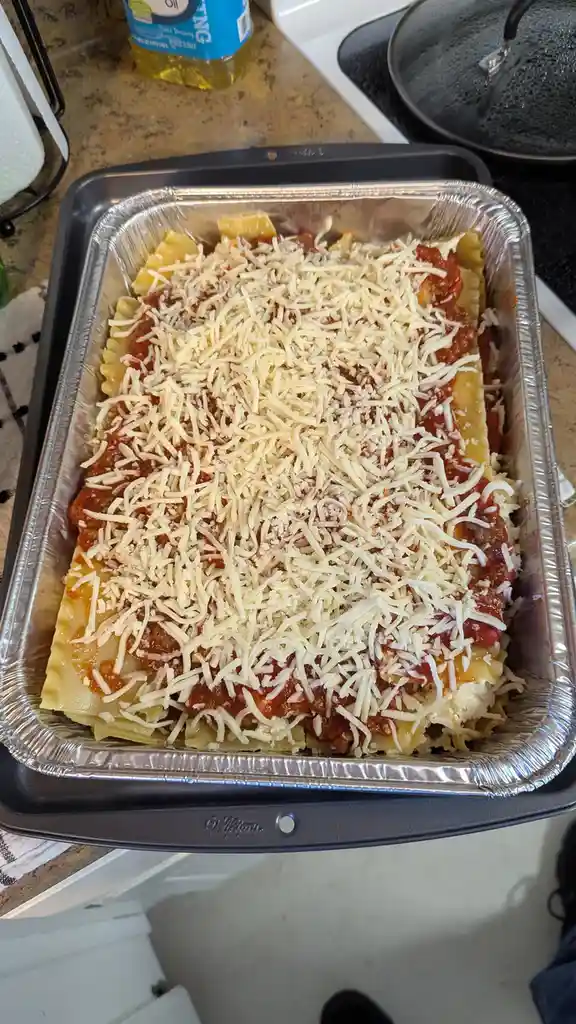 A close-up photo shows a rectangular aluminum baking dish filled with uncooked lasagna, featuring layers of pasta sheets…