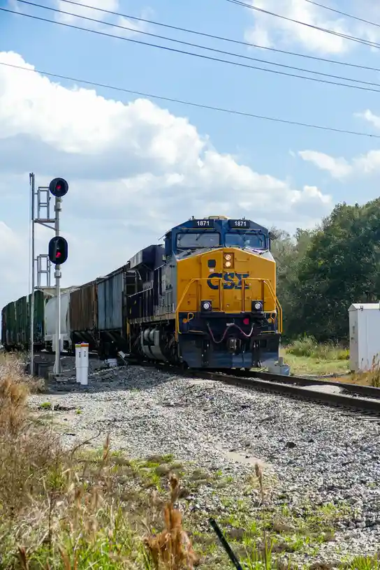 A CSX freight train, numbered 1871, approaches on a sunny day with a red signal light visible to the left. The overlaid text…