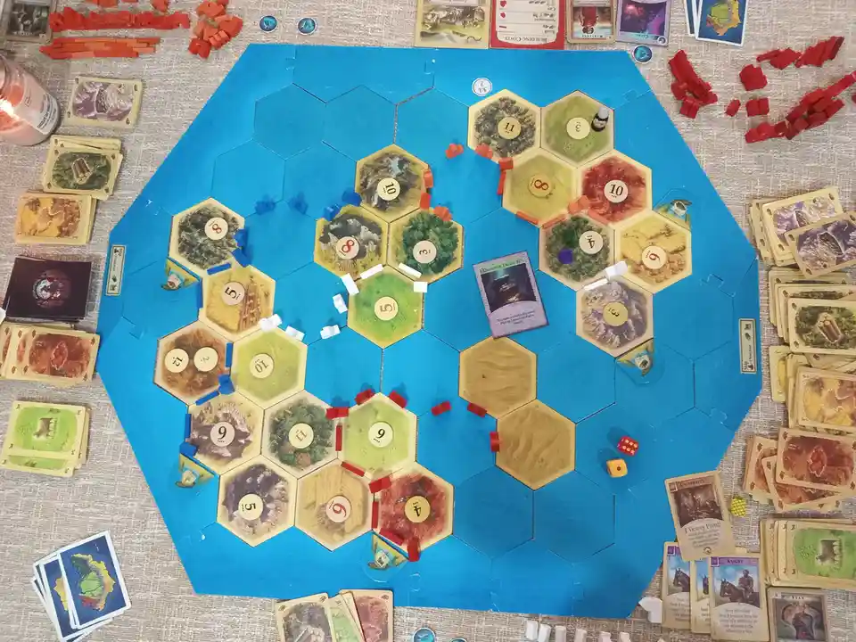 An overhead shot of a partially played board game with hexagonal tiles, resource cards, and game pieces scattered around…
