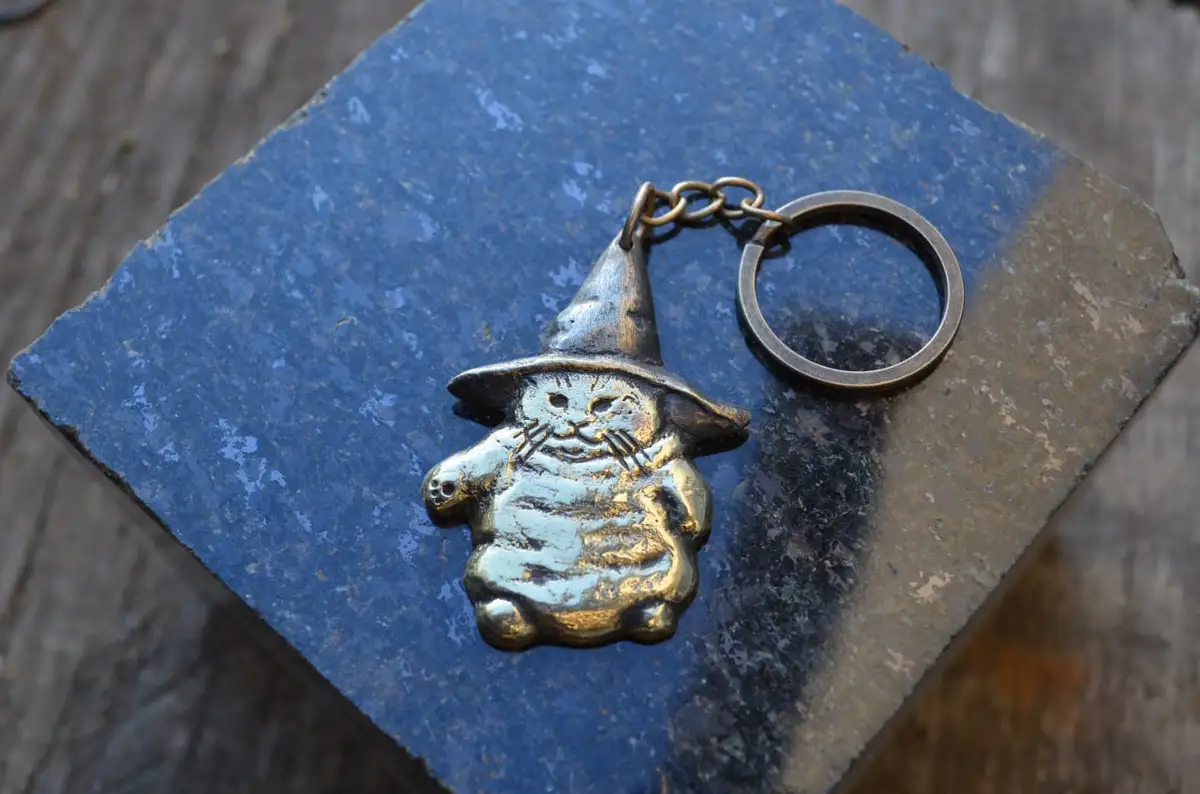 A small, metallic keychain shaped like a cat wearing a pointed wizard's hat rests on a blue, weathered stone surface. The…