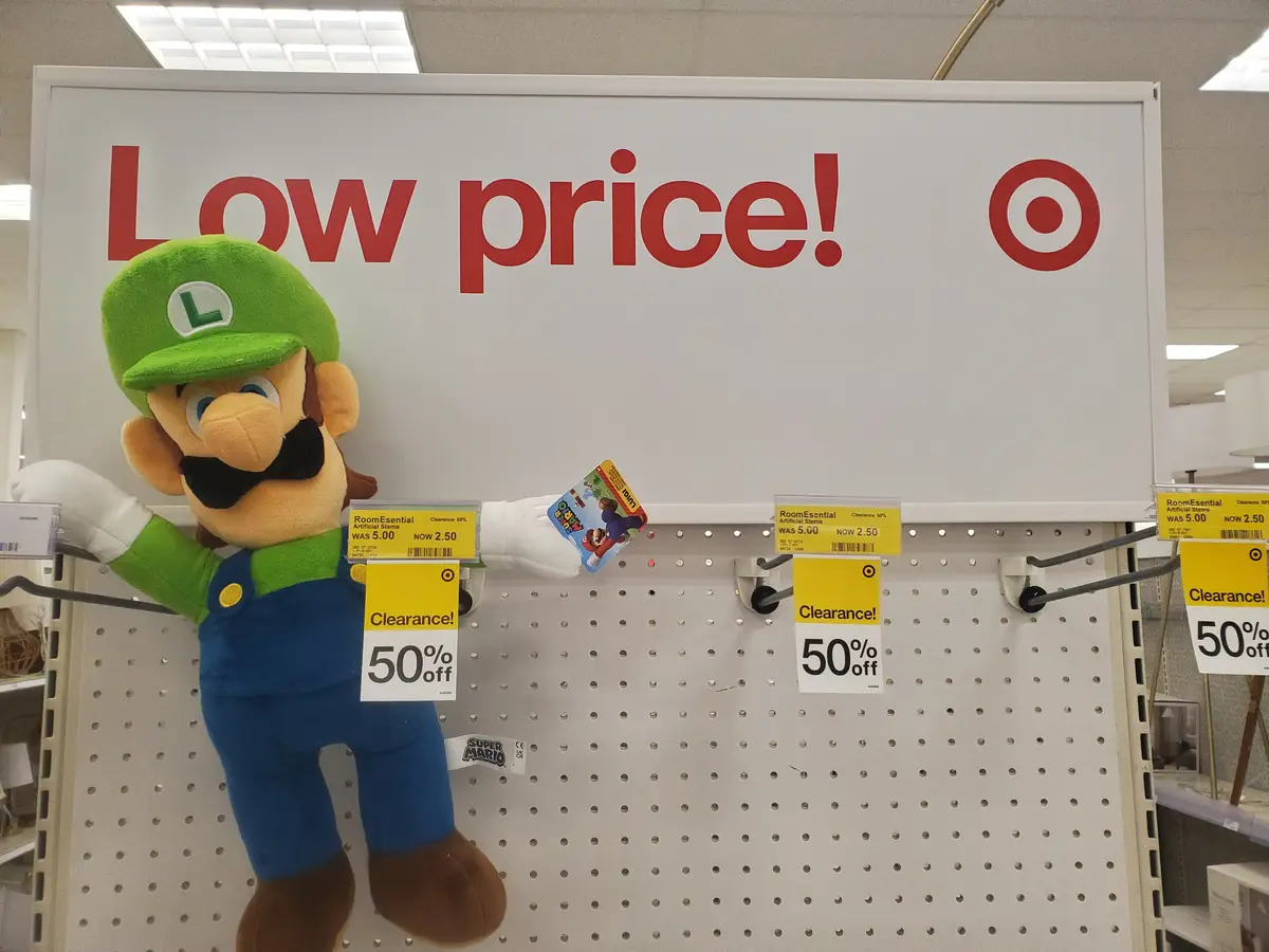 A large plush Luigi doll from Super Mario stands on a Target store shelf, holding a small Mario card, with multiple yellow…