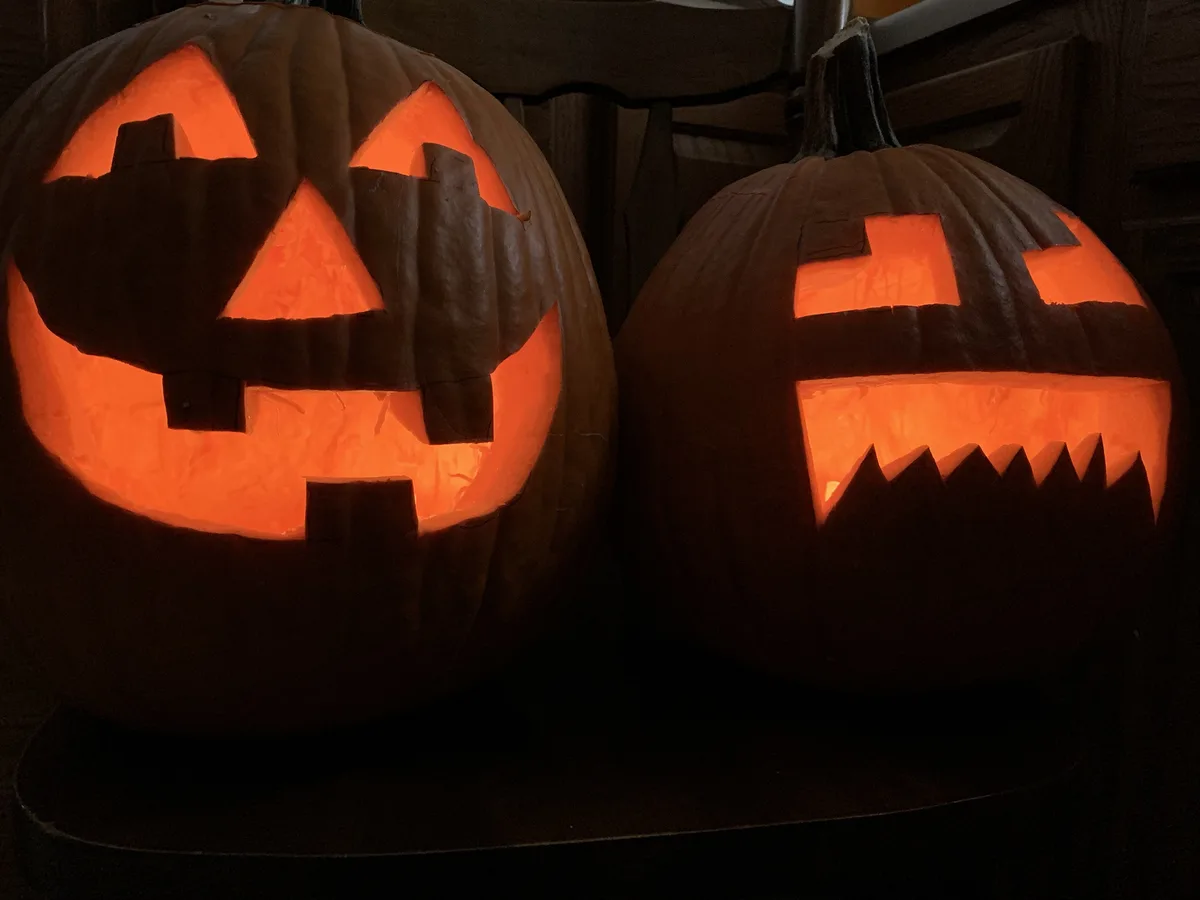 Two carved Halloween pumpkins glow with orange light in a dark room. The pumpkin on the left has a face with triangular…