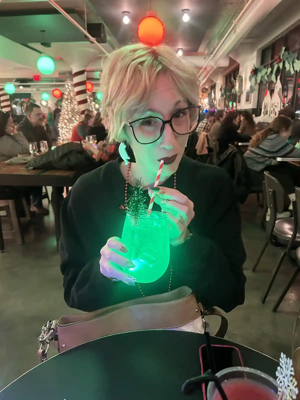 A person with short blonde hair and glasses is sitting in a festive, dimly lit bar decorated with Christmas lights and…