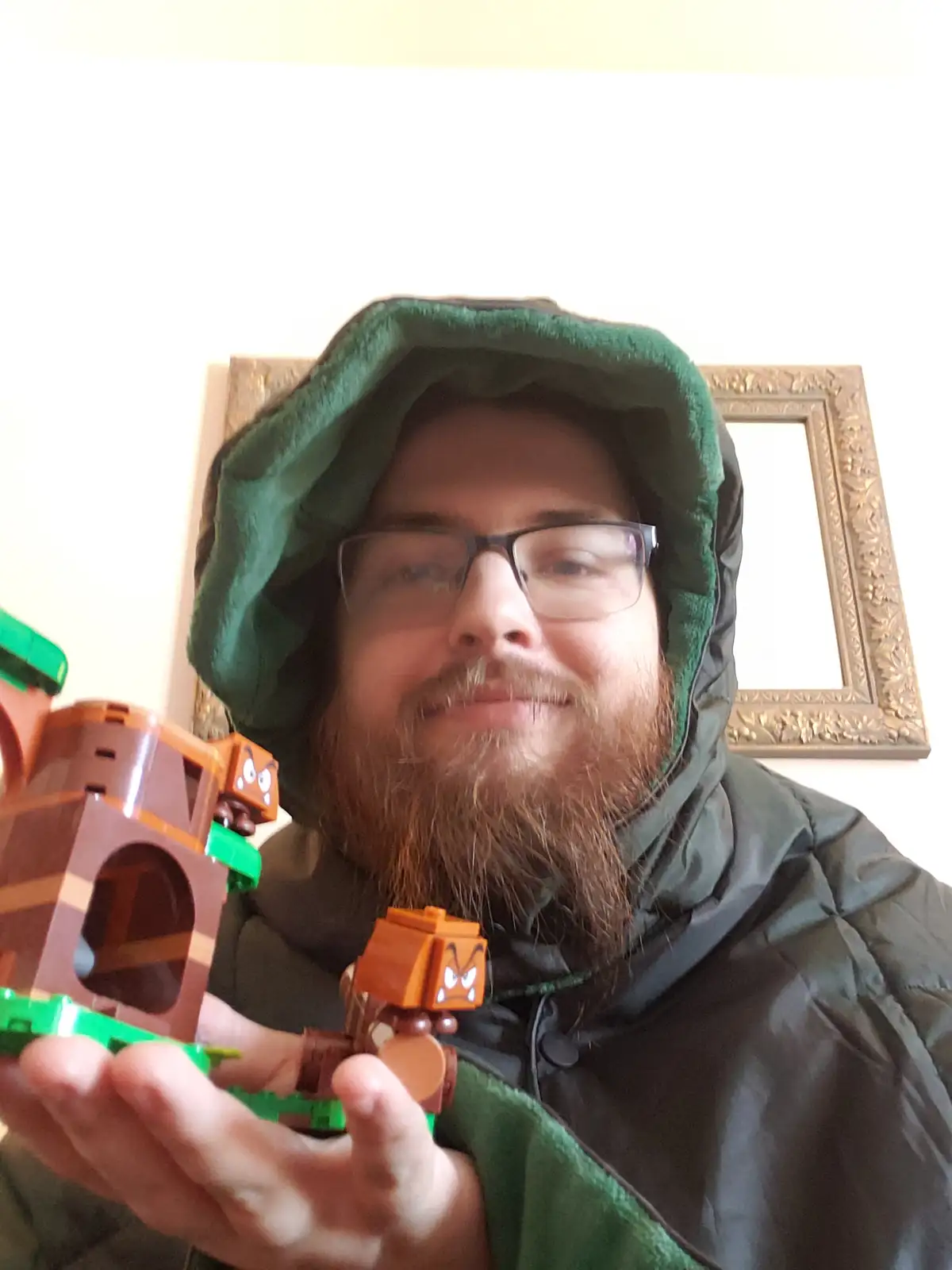 A man with a beard and glasses, wearing a green hooded jacket, holds a Lego Super Mario set featuring brown and green pieces…