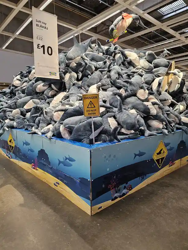 A massive pile of grey plush sharks fills a display at an IKEA store, with a yellow 'DANGER DO NOT ENTER' sign stuck in the…