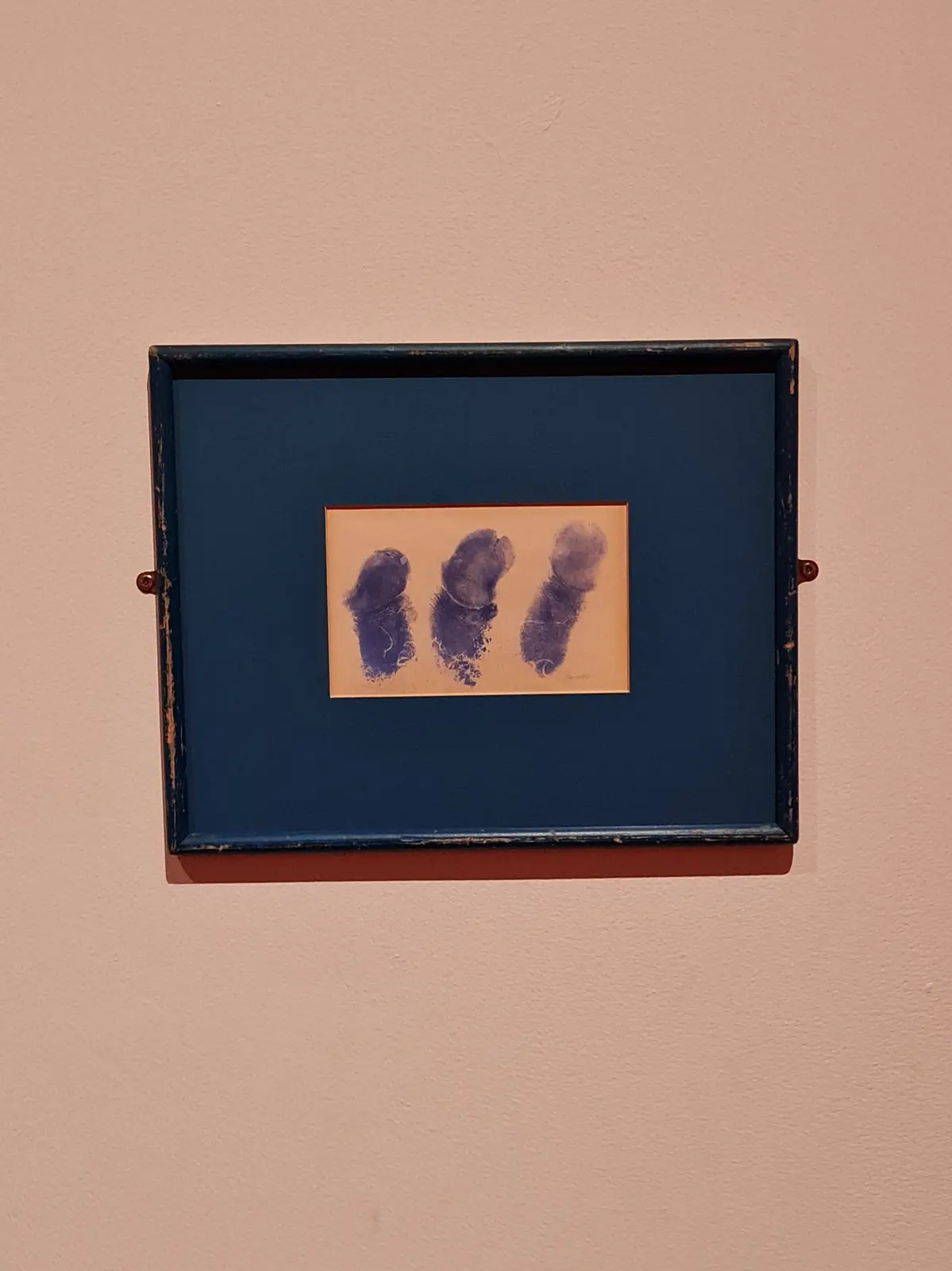 A framed artwork on a wall displays three dark blue fingerprints arranged horizontally on a beige paper, mounted within a…