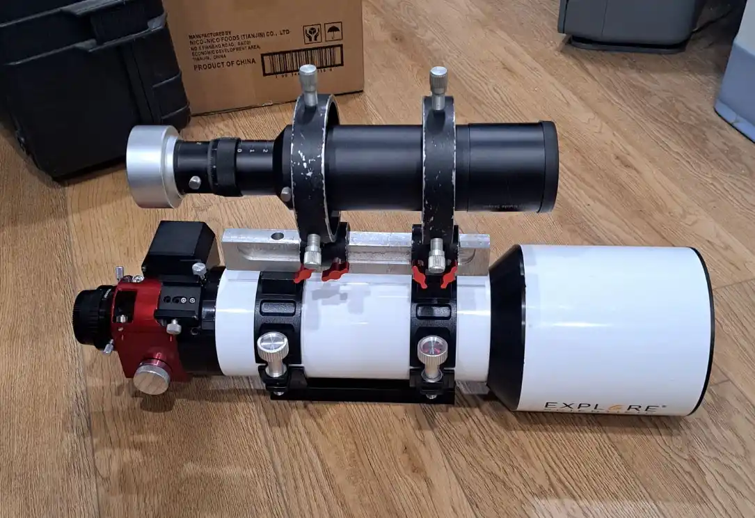 A white and black Explore Scientific telescope lies on a wooden floor, with a black camera mount and lens assembly attached…
