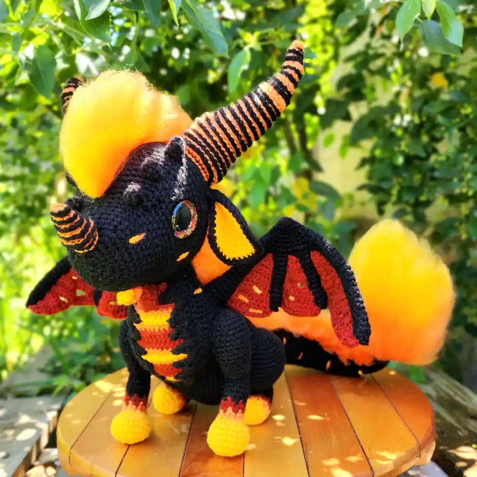 A handmade crocheted dragon plushie with black body, orange and yellow flame-like mane, tail, and feet, red and black wings…