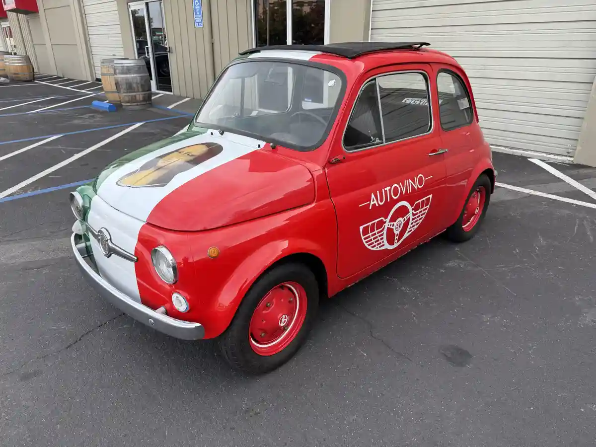 A vintage red, white, and green Fiat 500 car is parked in a lot, painted like the Italian flag with a portrait of a man on…