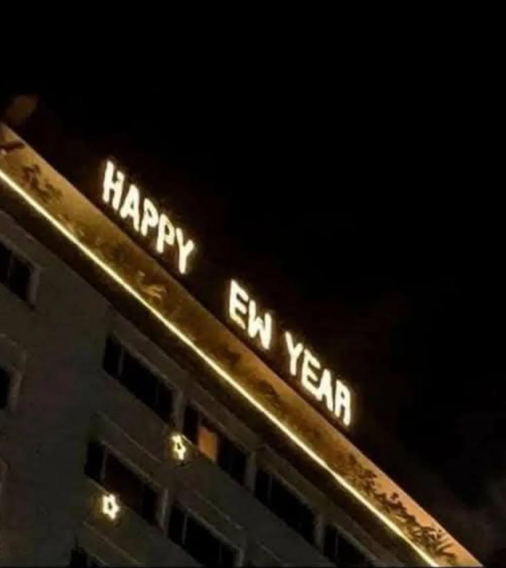 A nighttime photo of a building's illuminated sign that reads 'HAPPY EW YEAR', with 'EW' being a misspelling of 'NEW'. The…