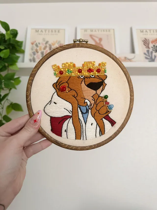 A hand holds up a circular embroidery hoop displaying a stitched cartoon of a brown bear wearing a jeweled crown, a white…