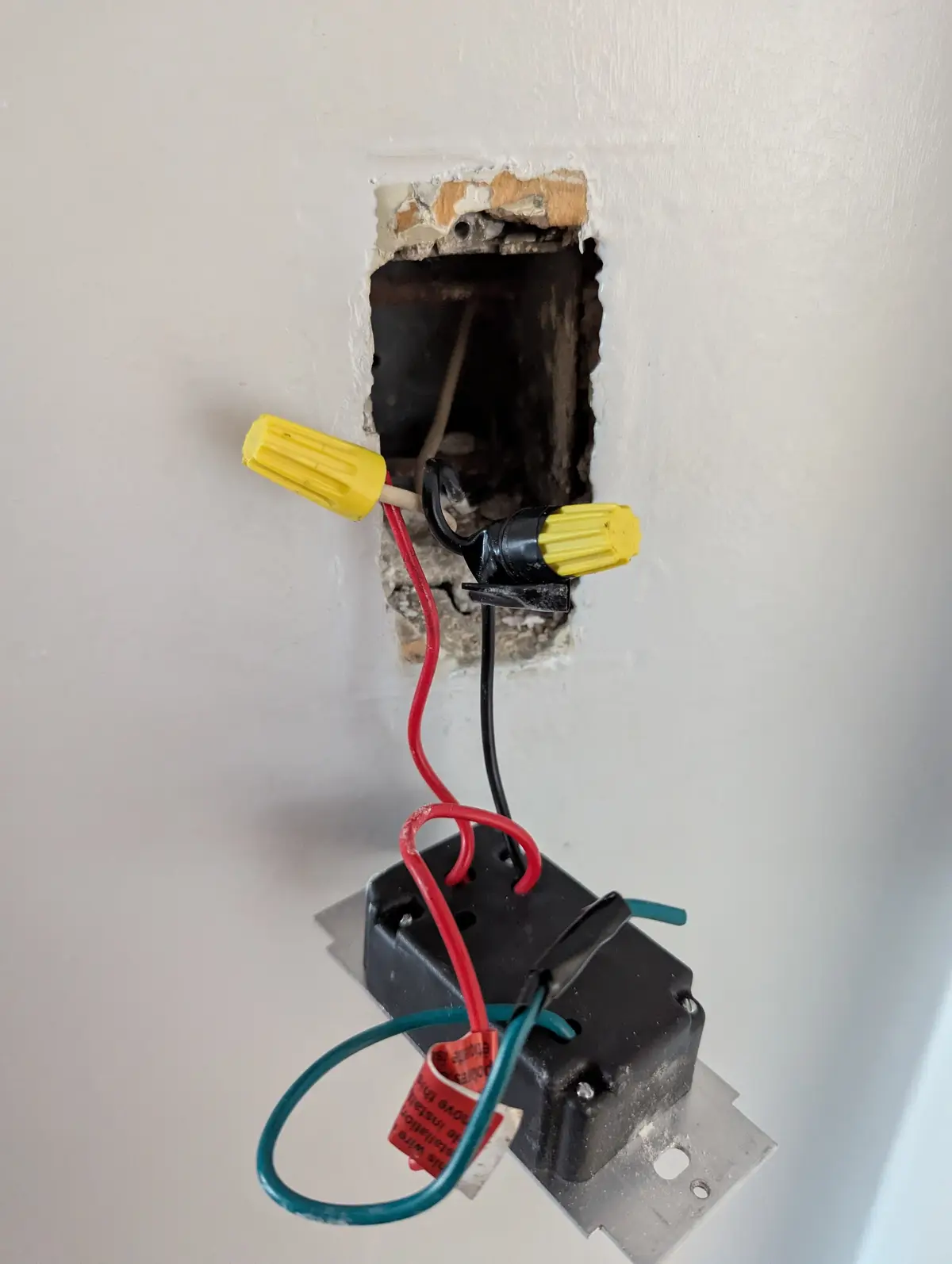A close-up photo shows an unfinished electrical installation in a white wall, with exposed red, black, and blue wires…