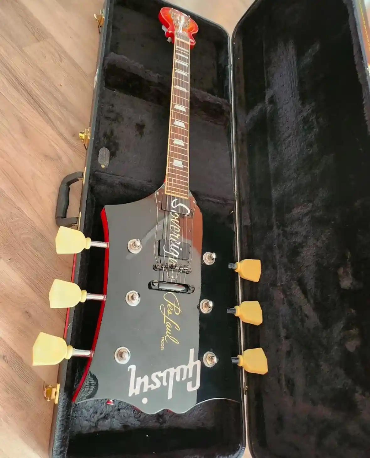 A black and red Gibson Sovereign Tex Faul Model electric guitar with yellow tuning pegs rests inside its open black…