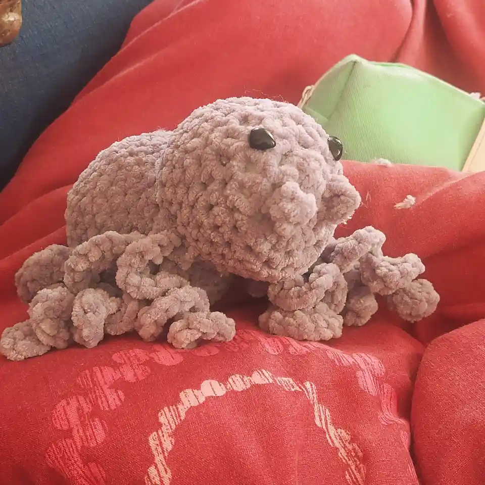 A handmade, light purple crocheted octopus with black button eyes sits on a red fabric surface, possibly a couch or blanket…