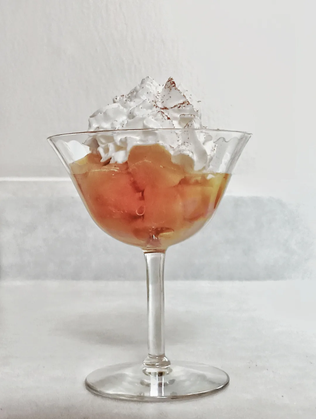 A dessert in a stemmed glass cup, featuring a layer of orange-colored fruit pieces in syrup, topped with a generous dollop…