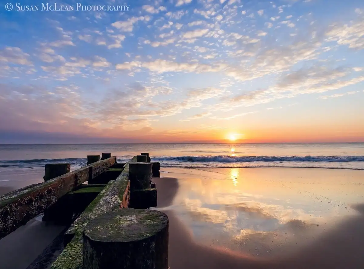 A serene beach scene at sunrise with a wooden pier extending into the water, reflecting the orange and blue sky. The…
