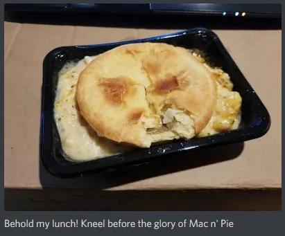 A close-up photo of a black takeout container holding a macaroni and cheese dish topped with a golden-brown, slightly…