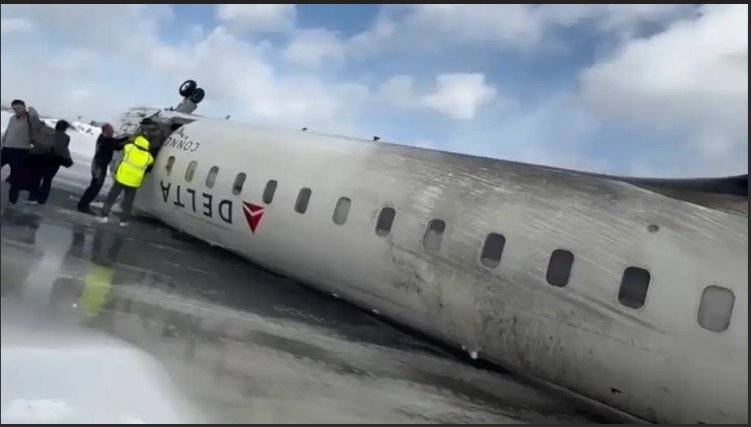 A Delta airplane fuselage lies on its side in a snowy field, with several people standing nearby, one in a bright yellow…