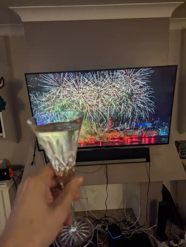 A person holds a crystal champagne flute filled with bubbly liquid in front of a TV displaying vibrant fireworks over a city…