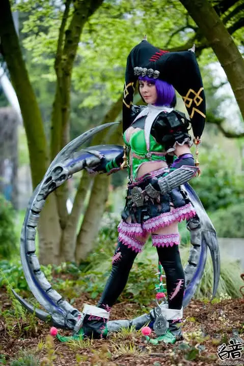 A person in a detailed cosplay of a fantasy character with purple hair, wielding a large scythe-like weapon, stands outdoors…