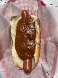 A hot dog in a bun is covered in a thick, dark brown sauce, sitting in a red and white checkered paper basket. The overlaid…